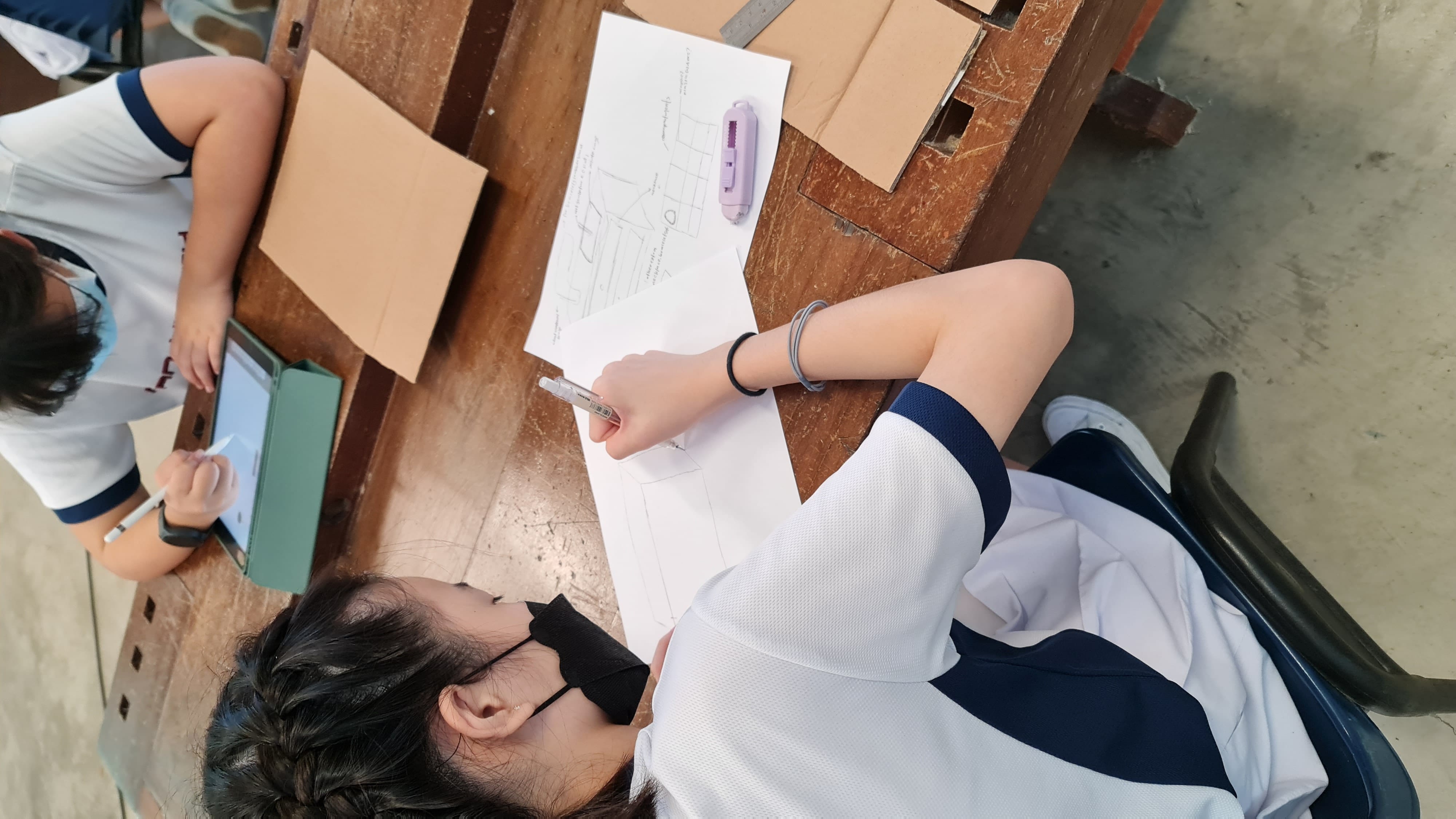 Two students in white uniforms sit at wooden desks; one draws on paper, the other writes on a tablet with a stylus.